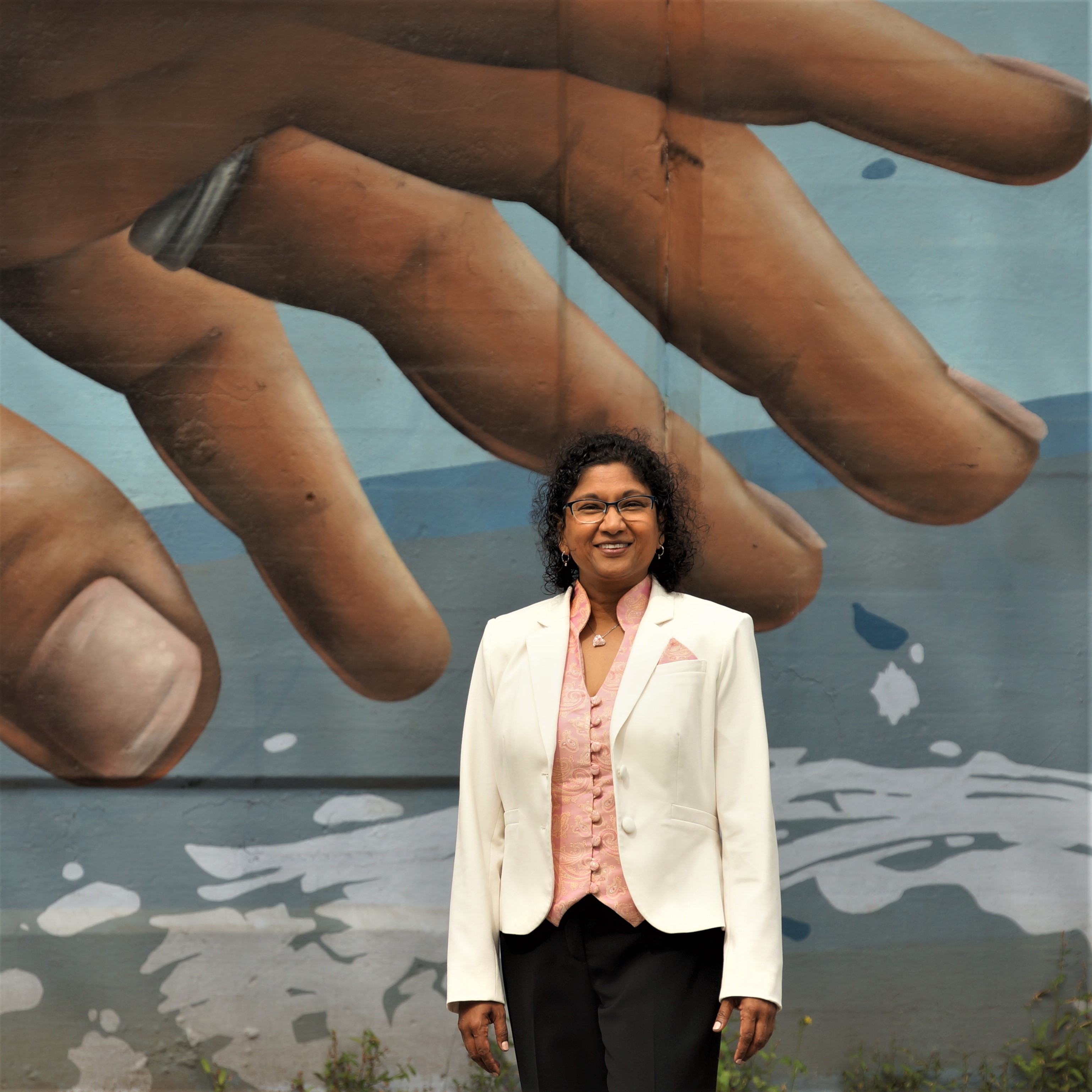 Wall mural of brown-skinned hand hovering over image of ME standing (ME = curly, black-haired, brown-skinned, smiling woman with glasses wearing a suit (white jacket, black pants and pink and gold paisley vest