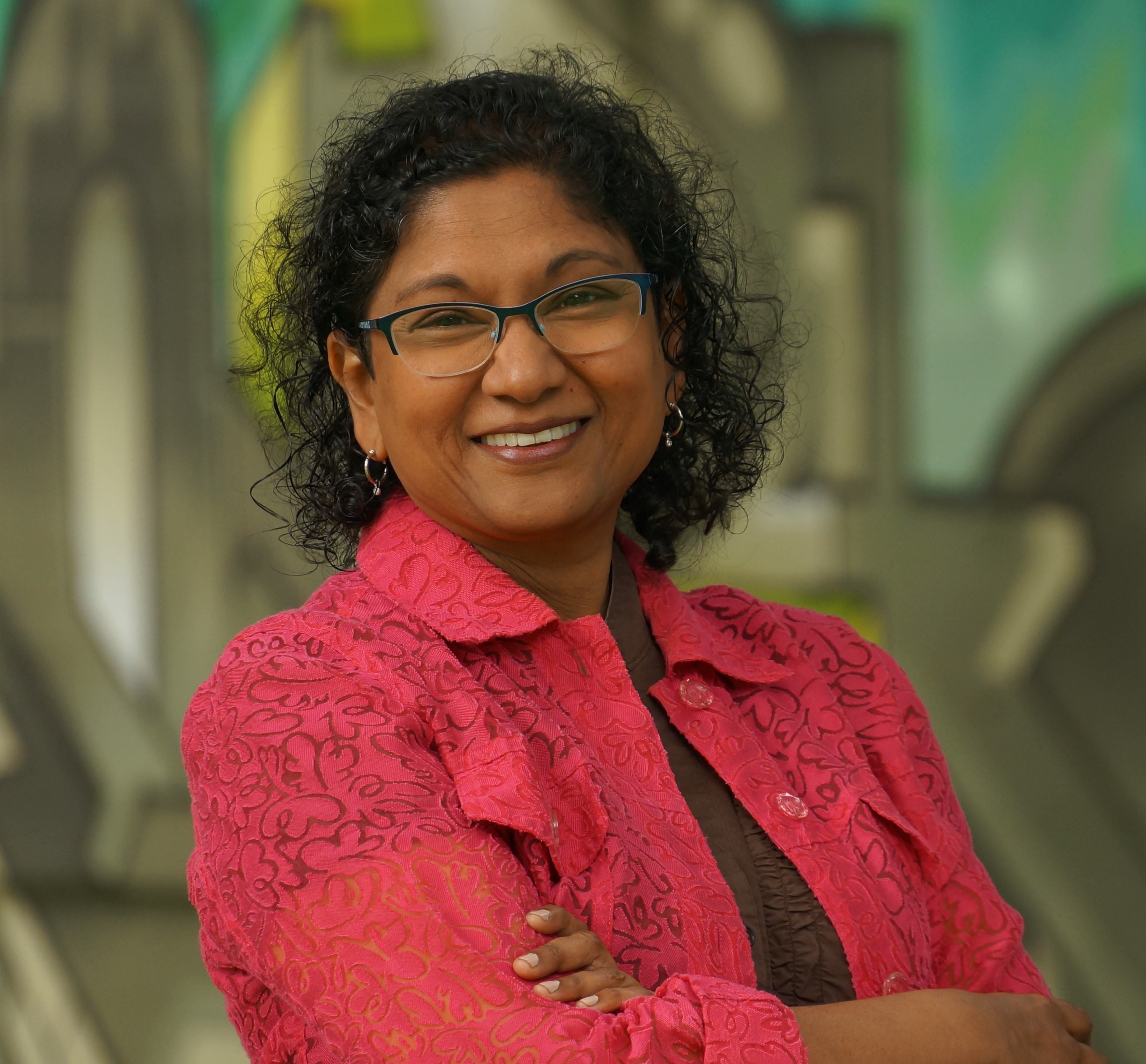 Head and shoulders photo of me (Me = curly, black-haired, brown-skinned woman wearing glasses, a pink laced jacket, brown shirt and a smile)